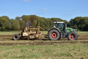 a green tractor pulling a yellow rotary ditch digger attachment in a field with mud furrows in the grass 